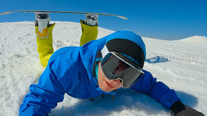 特写:一个年轻人在学习如何在滑雪板上转弯时跌倒在雪地上
