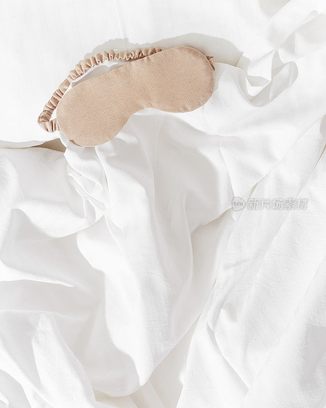 米色眼罩适合睡在家中白色的被褥上，极简的生活方式美学平躺照片。女性睡眠面膜，适合最佳睡眠者，适合旅行，舒适放松。好好休息的概念。