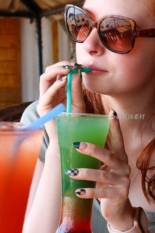 特写照片中，迷人的红发女子用塑料吸管、冰块喝着彩虹模拟鸡尾酒，在户外戴着太阳镜，桌子上放着龙舌兰日出鸡尾酒，露天用餐背景