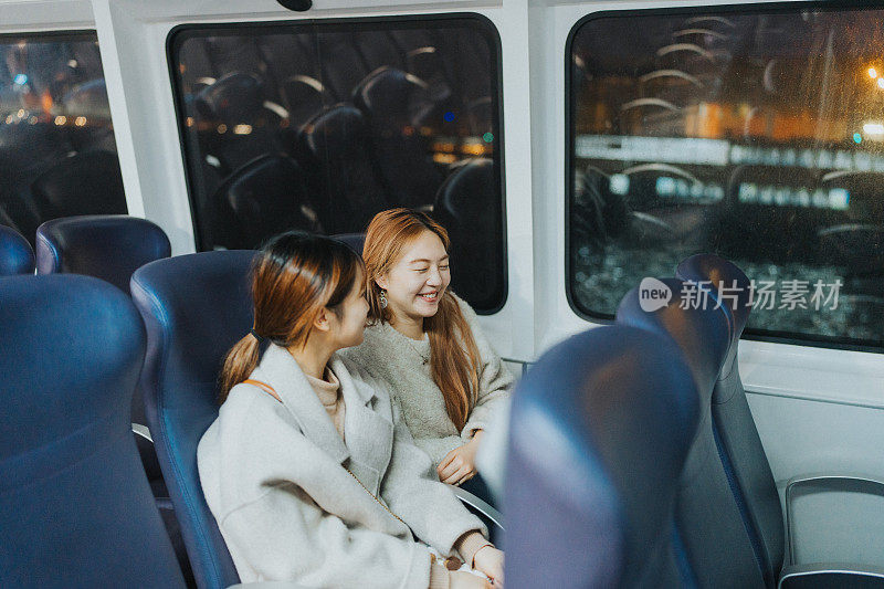 两名年轻的亚洲女子坐在香港的一艘渡轮内，夜幕降临时，背景是城市的灯光。