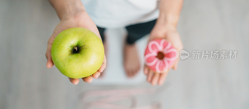 体重秤上的女人手里拿着青苹果和甜甜圈，选择水果是健康的，而甜食是不健康的垃圾食品。节食控制，体重减轻，肥胖，饮食方式和营养