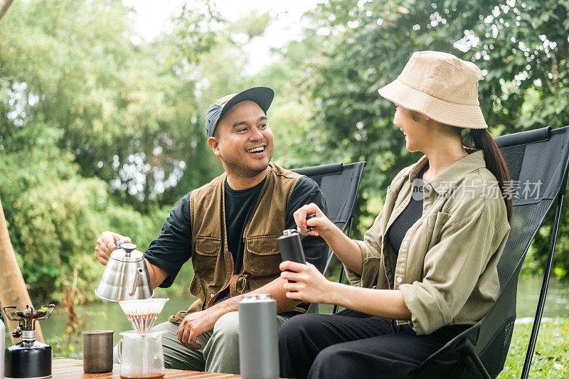 年轻的亚洲夫妇，男人和女人坐在户外露营地喝咖啡。两个人带着咖啡机工具在森林里露营。旅游，放松，露营，度假，假日，周末的主题