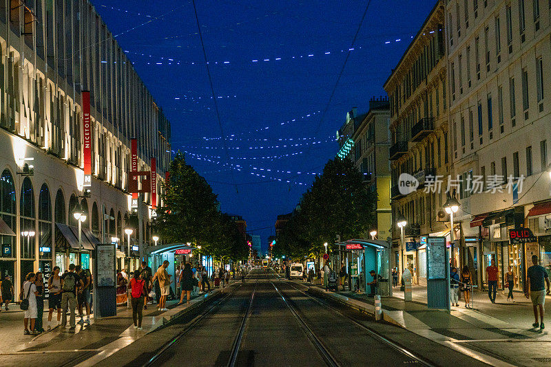 在法国里维埃拉的尼斯，黄昏中的让·梅迪辛，在电车站行走和等待的人们，照亮城市的街灯。