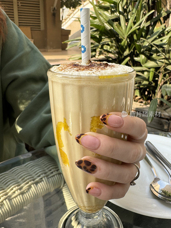 特写图片:一个无法辨认的年轻女子手持香蕉奶昔和纸吸管，猎豹动物印花指甲艺术，在露天咖啡馆餐厅花园饮用无酒精饮料，重点放在前景