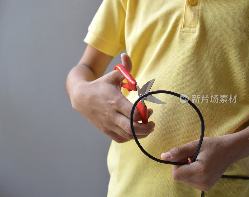 一只手拿着一圈电线或充电线，在黄色t恤和灰色背景上用红钢剪刀剪断