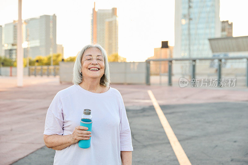 在运动场上，一位快乐的年长妇女站在那里，手里拿着水瓶，面带微笑