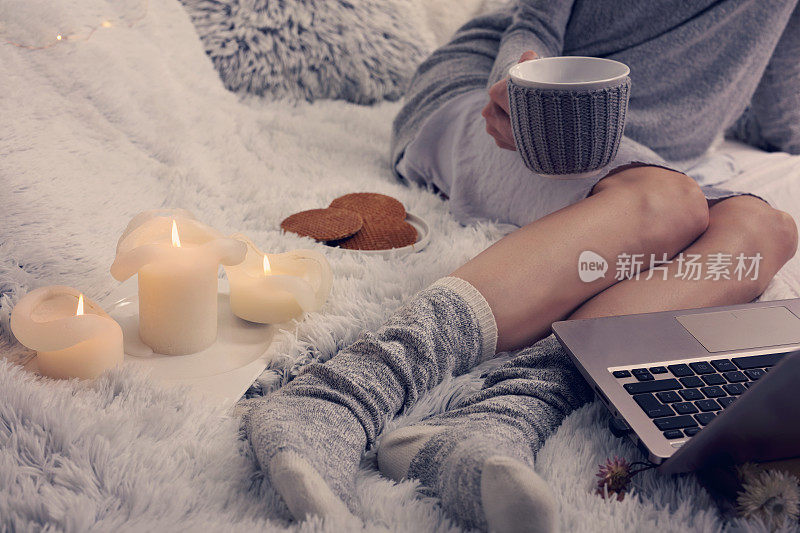 舒适的夜晚，温暖的羊毛袜，柔软的毯子，蜡烛。女人在家里放松，喝着可可，用着笔记本电脑。舒适的生活方式。