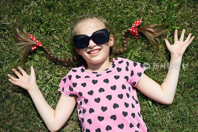 女孩戴着太阳镜躺在绿色的草地上，穿着粉红色的衣服带着爱心，阳光明媚，夏日户外，俯视图