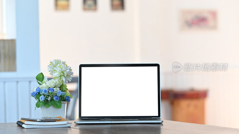 白色的空白屏幕笔记本电脑，笔记本电脑，智能手机和盆栽植物，所有这些都放在现代的办公桌上，以舒适的客厅为背景。