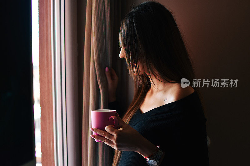 年轻漂亮的女人在早上透过窗户看和喝咖啡