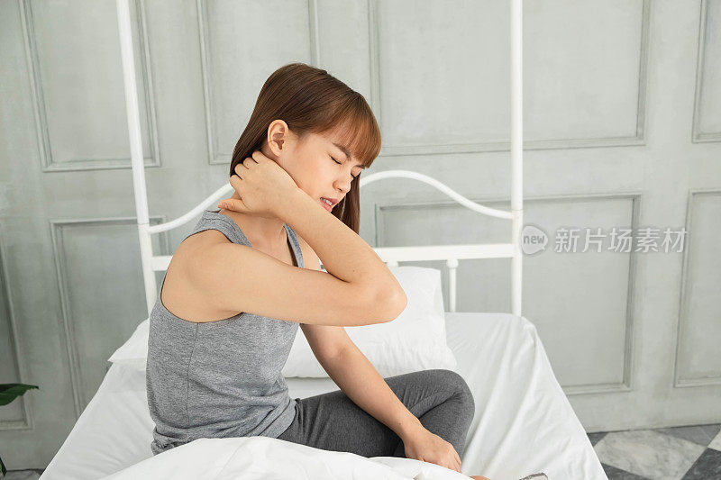 年轻美丽的亚洲女人穿着灰色的衣服，早上醒来在白色的床上，身体疼痛，拥抱的手在腹部蜷缩，腹痛，腹痛，痛，痛