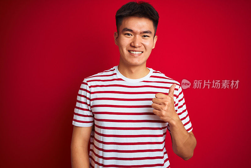 年轻的亚洲华人男子穿着条纹t恤站在孤立的红色背景做快乐的拇指手势与手。赞许的表情看着镜头，表示成功。