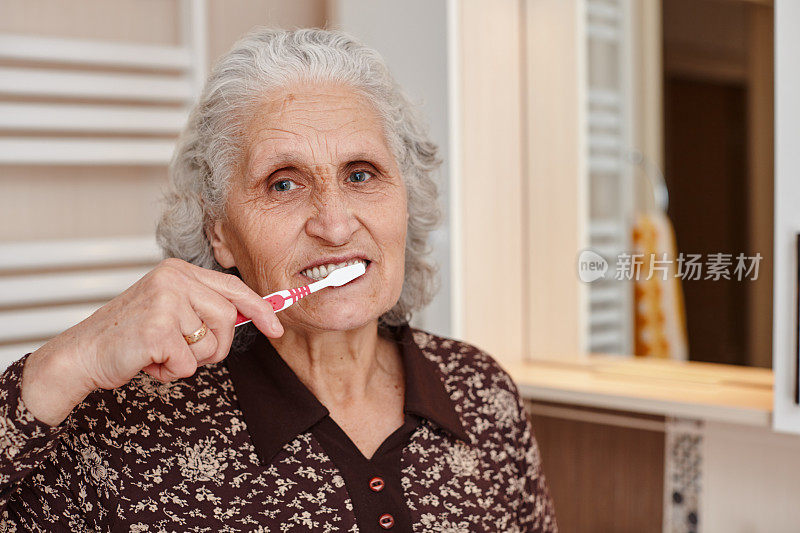 美丽的老妇人与灰色的头发刷牙，同时看着镜头在浴室的镜子和水槽