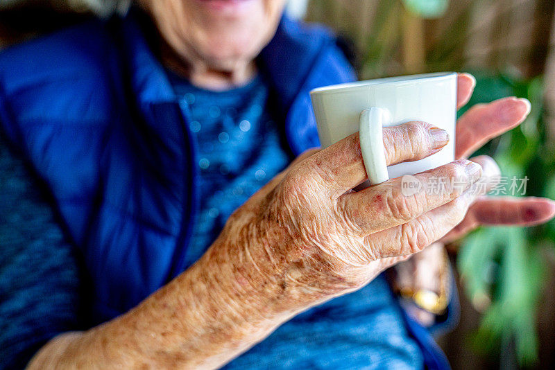 近距离拍摄的老年人白种人的手拿着一个白色陶瓷杯里的咖啡或茶在室内看窗外在夏天