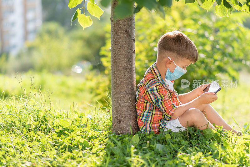一个戴着医用口罩的孩子坐在草地上，看着夏日日落时的手机卡通。手里拿着手机的孩子。大流行期间的防疫工作