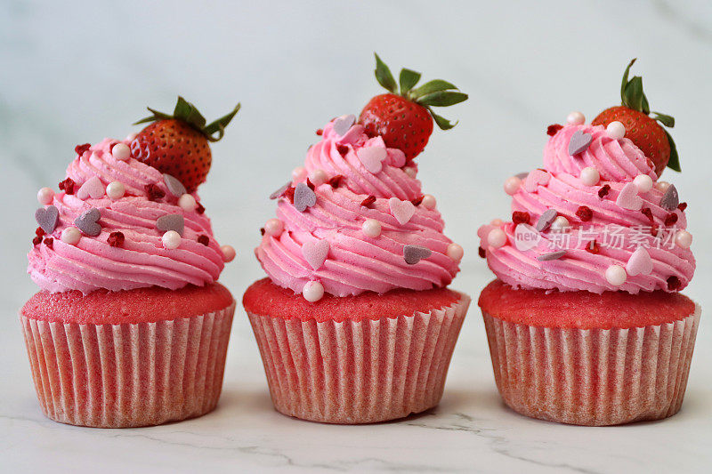 近距离图片三排，自制，粉色天鹅绒草莓纸杯蛋糕在纸蛋糕盒，草莓上的粉色奶油糖霜管道漩涡，糖工艺心形和球形点缀，在大理石蛋糕架上，关注前景