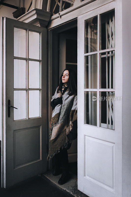 一个年轻的白人女孩站在门口，穿着秋天的深色外套，围着宽大的围巾，戴着手套，望着远方。有玻璃的旧门。