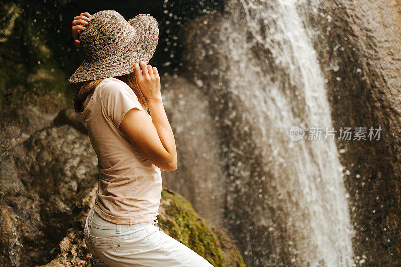 一个美丽的年轻女子，头上戴着一顶帽子，站在瀑布旁边
