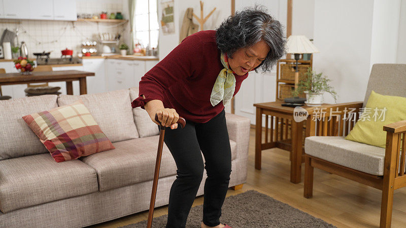 背部疼痛的亚洲受伤老年女性正拄着拐杖慢慢走向沙发，坐下来休息。