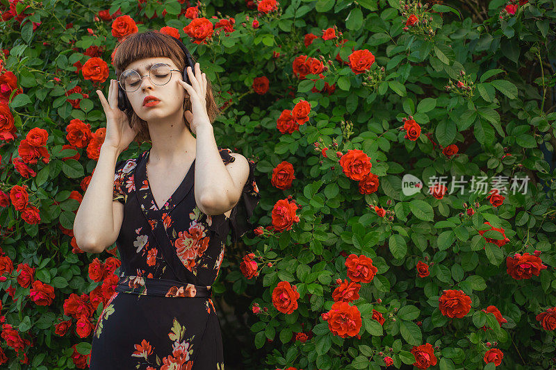 红头发的模特在玫瑰丛的背景下听音乐。亚洲女孩的穿着享受旋律。玫瑰花园。女人肖像。春天的花。鲍勃发型。愉快的声音。眼镜。学生
