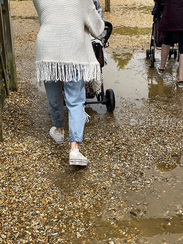难以辨认的人推着婴儿车通过水坑砾石路的图像，后方的观点，集中在前景