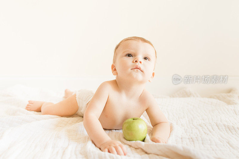 2022年秋天，一个9个月大的大流行男婴穿着可持续制作的棕色针织紧身衣，在一个新鲜的绿色史密斯奶奶苹果上玩耍和长牙，坐在家里地板上的奶油色薄纱毯子上