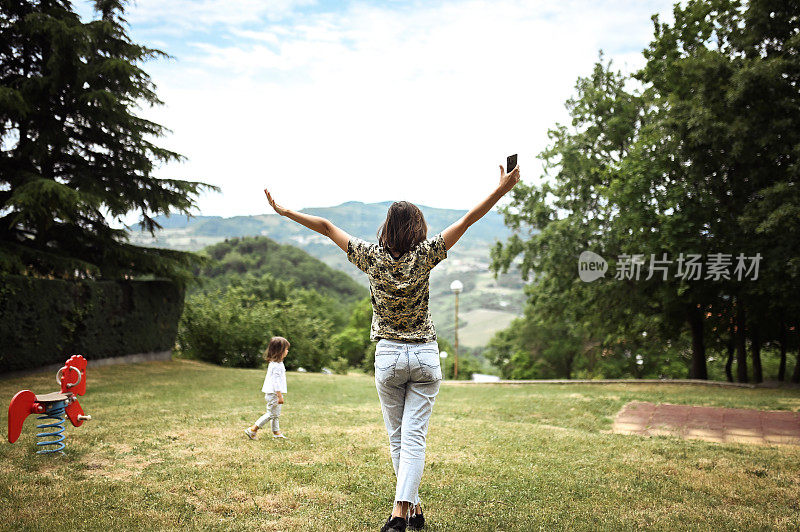 自由快乐的妇女与手臂放松在公园和她的孩子在操场上玩。