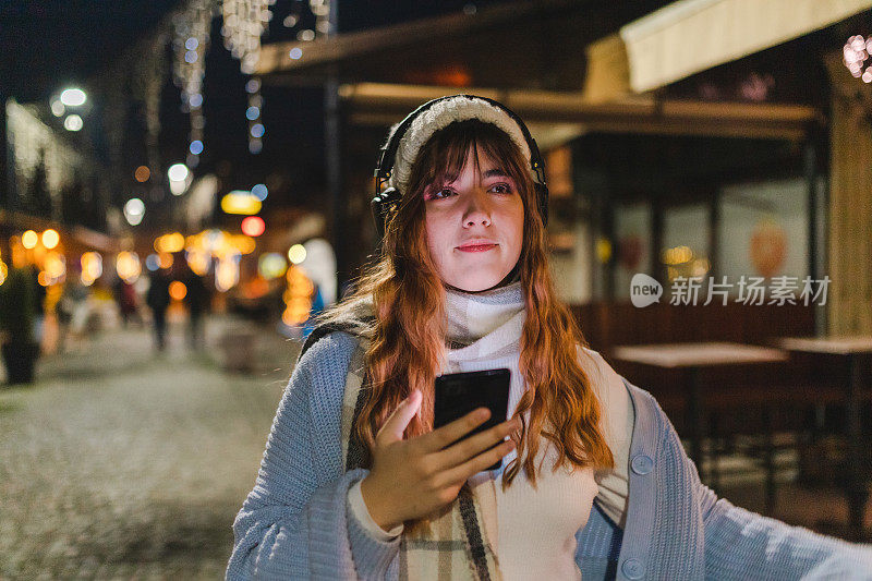 在城市的夜晚，微笑的年轻女子用她的智能手机听音乐