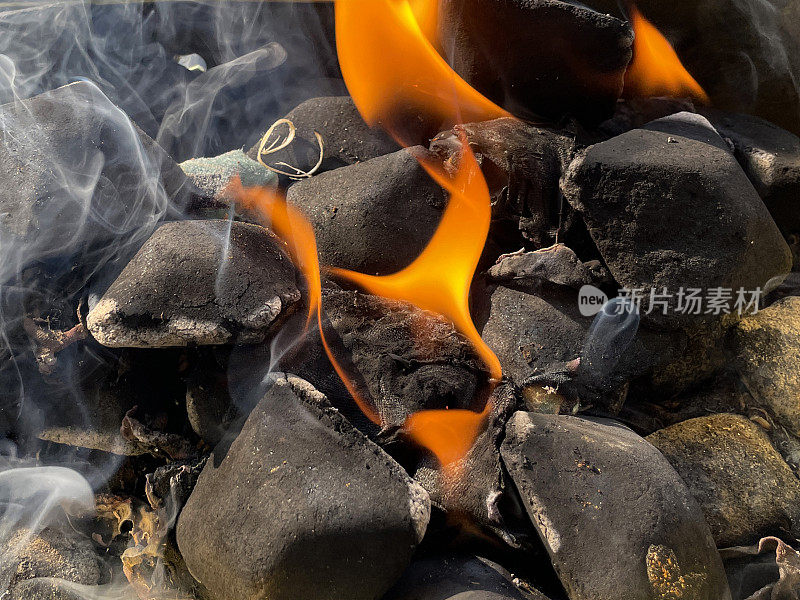 一个充满了燃烧的煤炭和木材的火坑的图像。在一个长方形烧烤架的黑暗中，木炭在火焰旁发光，近距离，高架视图