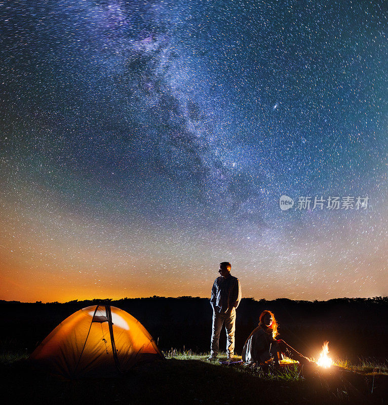 一对浪漫的徒步旅行者在他的营地在晚上靠近篝火和帐篷对星空与银河