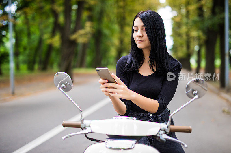 微笑的年轻女子看着她的手机，坐在摩托车上