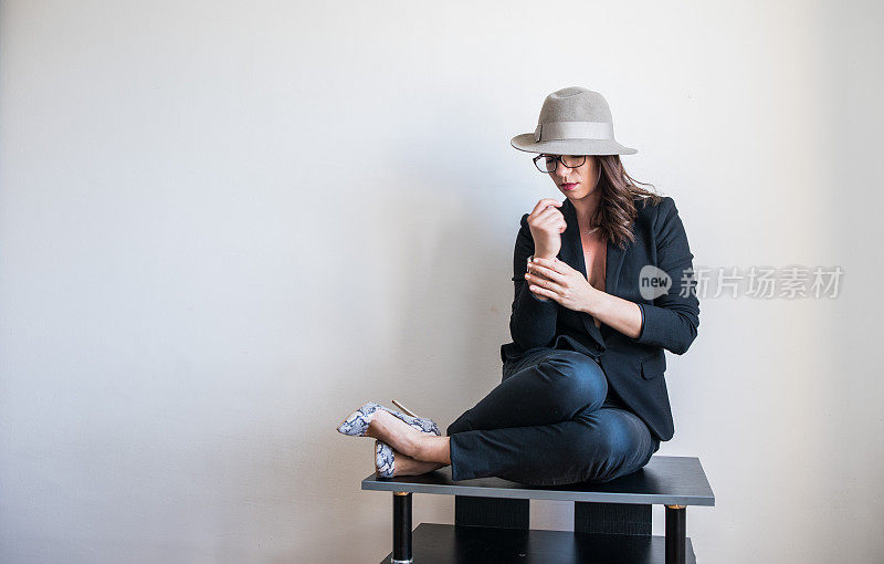 戴着眼镜和帽子的漂亮女孩坐在一张小桌子上，在一堵白墙前摆姿势