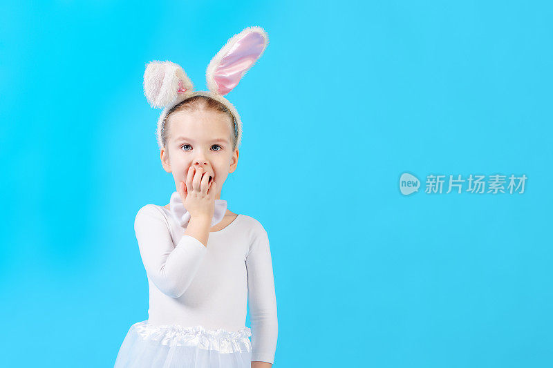 一个在蓝色背景下穿着白兔服装的孩子。可爱的小女孩长着野兔的耳朵，惊讶地捂着嘴。广告图片与文字空间。