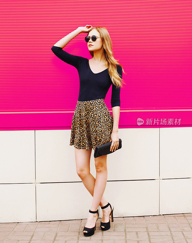 时尚的年轻金发女人在豹纹裙，太阳镜与手袋手袋手袋在城市街道上摆出彩色的粉红色墙壁背景