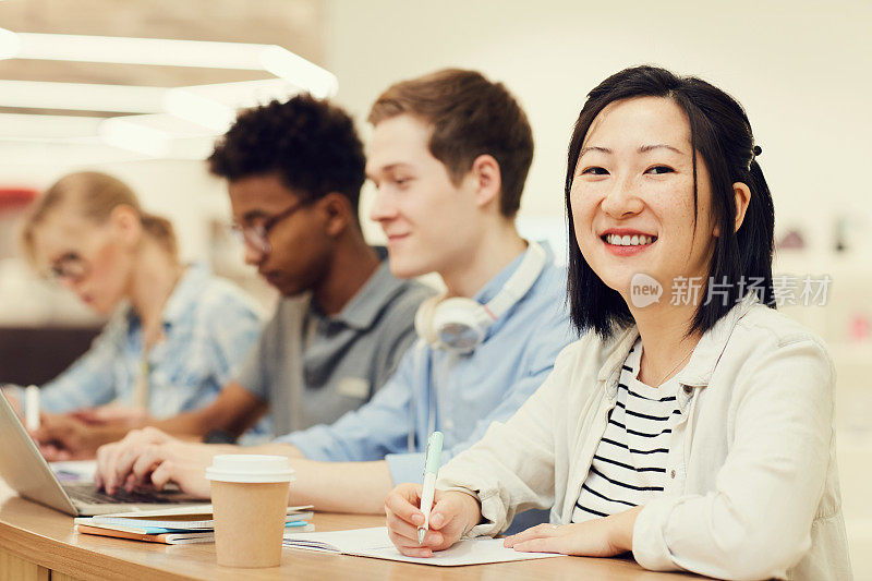 微笑的年轻亚洲女学生的肖像与短头发坐在桌子上与同学在讲堂和总结信息