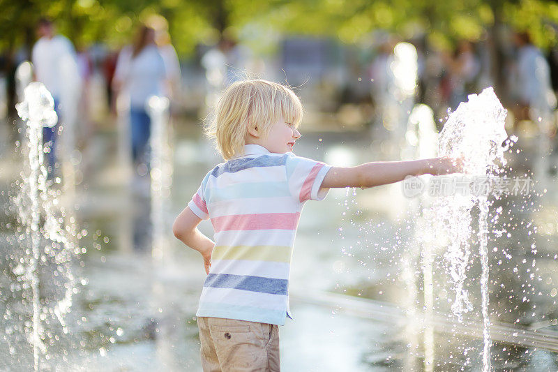 在阳光明媚的夏日，小男孩在广场喷泉的喷水口之间玩耍。活跃的夏季休闲城市的孩子。