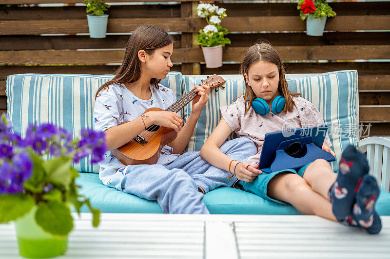 两个女孩拿着四弦琴、耳机和平板电脑在阳台上