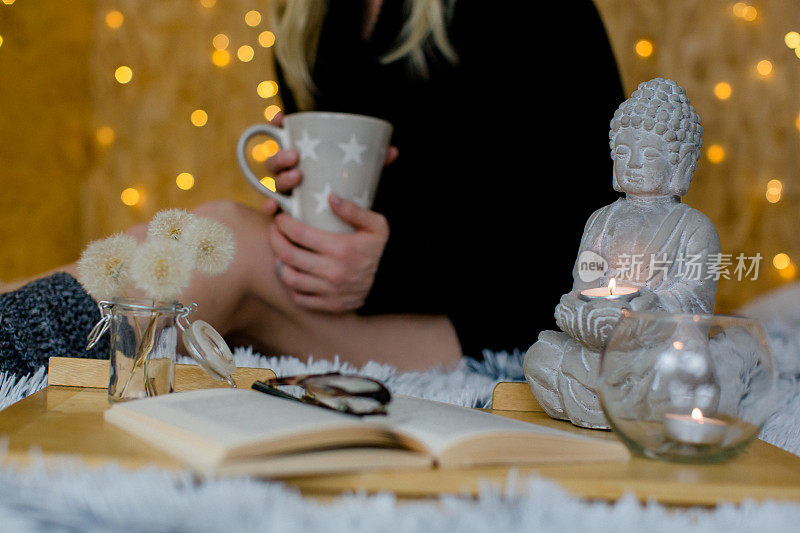 佛像，蜡烛和一本打开的书在前景-女人正在喝茶休息