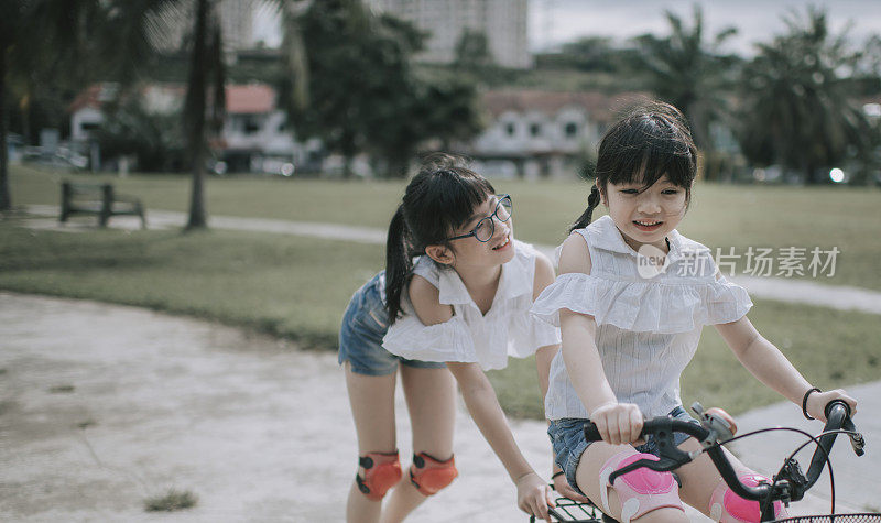 周末早上，亚洲华人姐姐在公园里推着她的妹妹学骑自行车