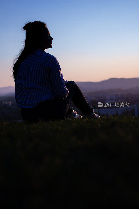 一个女人的黑色剪影坐在草山上俯瞰风景。