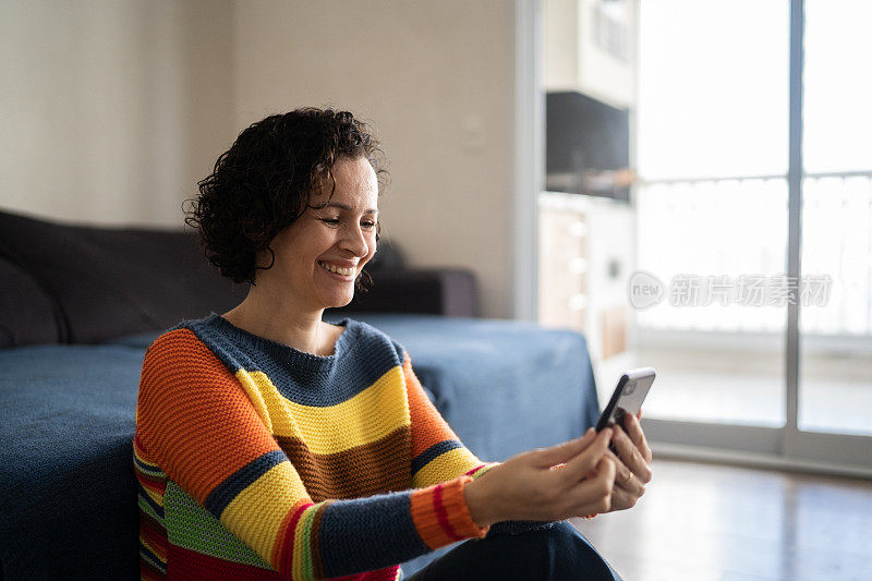 成熟的女人坐在家里的地板上玩智能手机