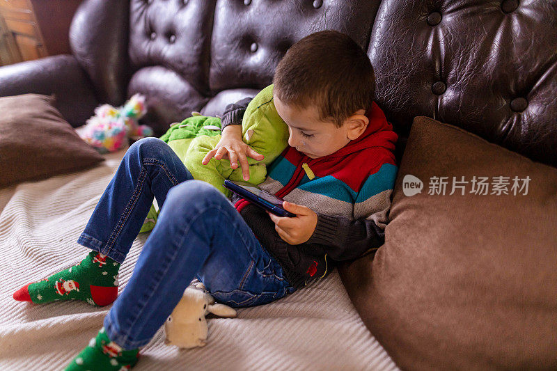 一个白人男孩一边抱着他的恐龙玩具一边用手机玩电子游戏
