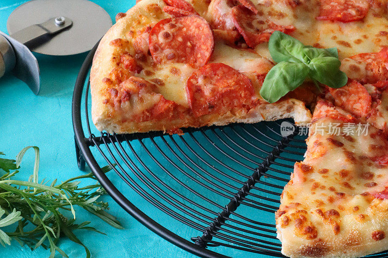 切成薄片的意大利辣香肠披萨在圆形冷却架上的特写图像，披萨轮，火箭叶，蓝色背景，前景聚焦