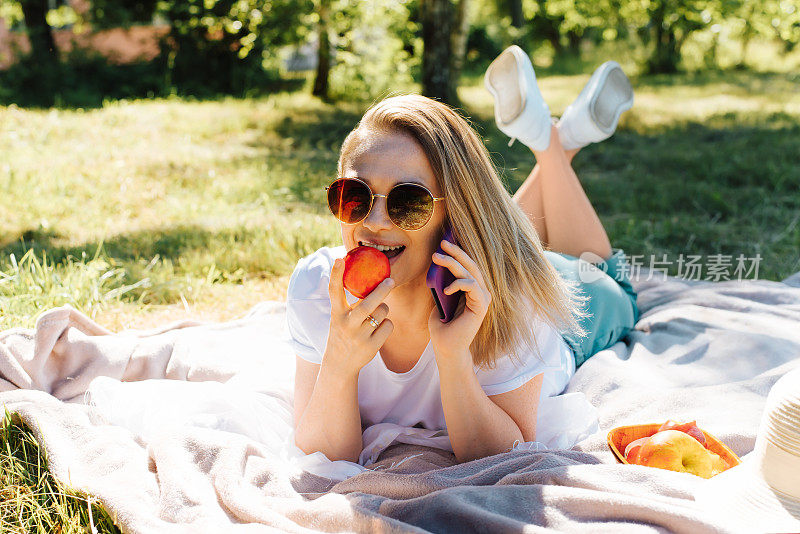 一个放松的年轻女人的肖像，戴着眼镜躺在草地上的毯子上，吃水果，同时打电话。美丽快乐的女孩在大自然中野餐休息。