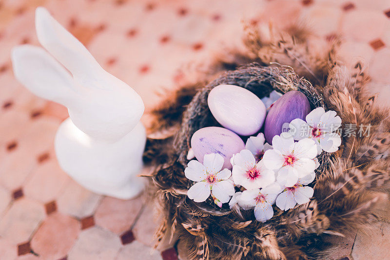 白色的复活节兔子，棕色的羽毛窝，紫色的复活节蛋和杏仁花放在一张质朴的户外石桌上