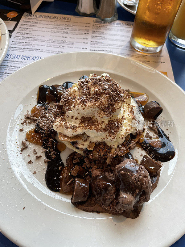 美式巧克力煎饼，奶油，巧克力酱，巧克力块和磨碎的巧克力放在白色盘子上，餐厅桌子背景，高视图，重点在前景