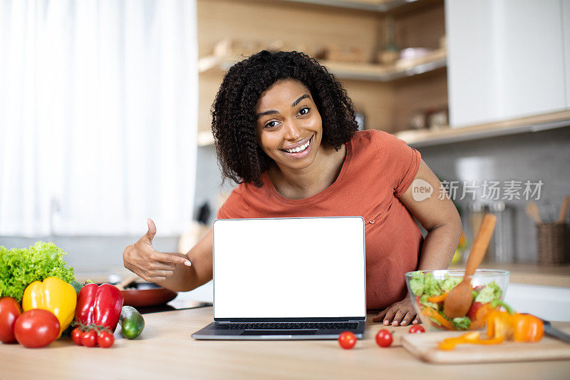 快乐的年轻非裔美国女士显示笔记本电脑与空白屏幕在餐桌与有机蔬菜在厨房