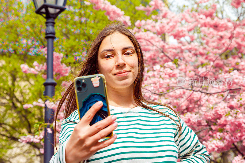 女子用手机视频通话，自拍。年轻微笑的欧洲白人女子在樱花樱花的背景，户外公园。