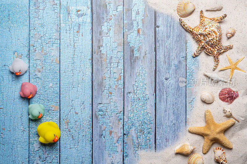 夏天，海滩和度假的概念与自由文本空间。前视图。平面布局，一小群彩色橡皮鸭，大海星和海贝壳和各种海贝壳和细沙滩在一个古老的蓝色木板背景