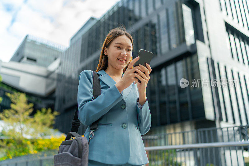 移动中的商业。连接移动。一位年轻的亚洲女商人背着笔记本电脑包，用智能手机上下班，她在中央商务区上班，对面是公司大楼